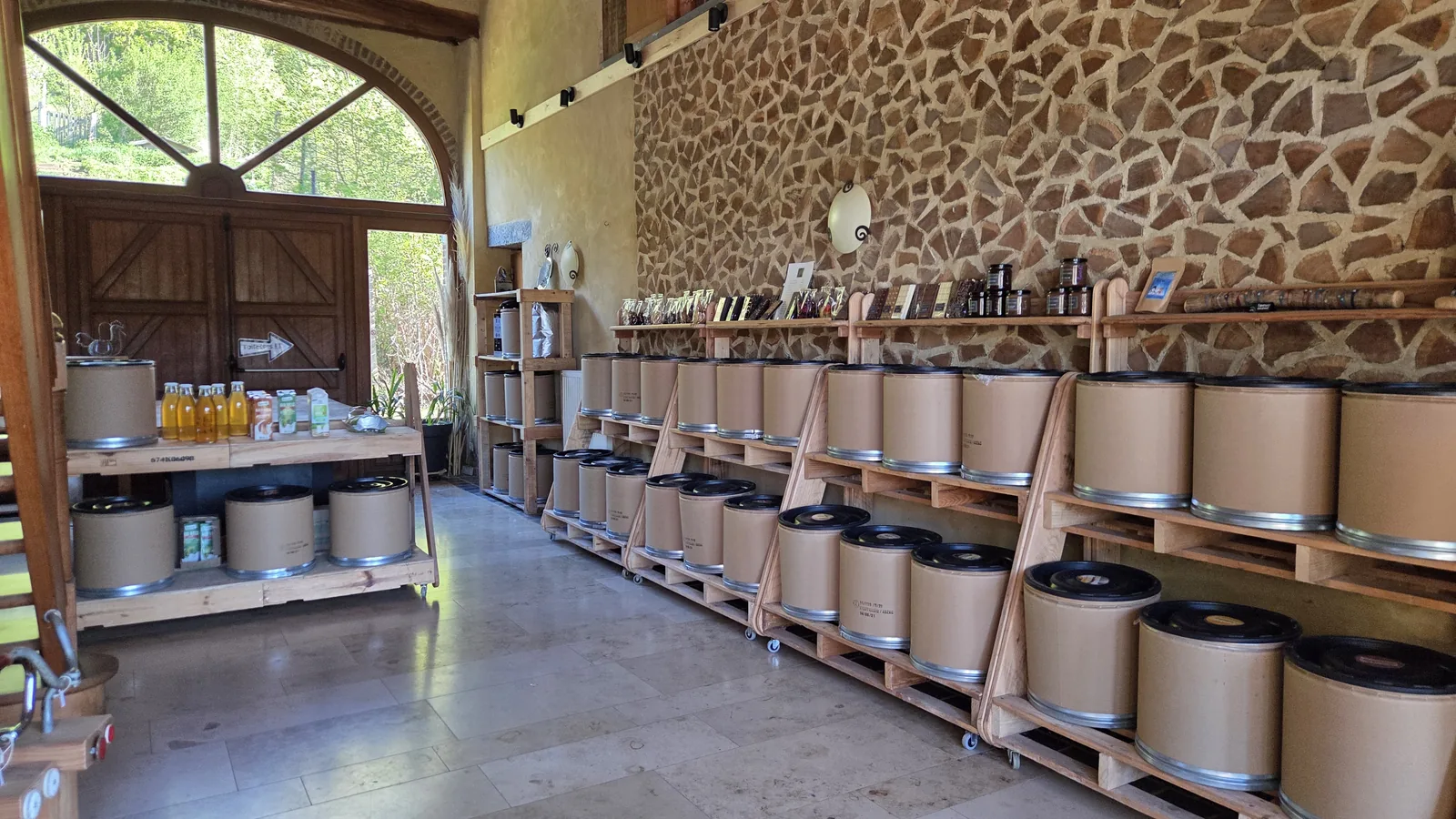 Intérieur de l'épicerie en vrac, une longue arche en pierre, plusieurs rangées de gros bidons en carton avec couvercles métalliques posés sur des étagères en bois, lumière naturelle qui entre par une grande baie