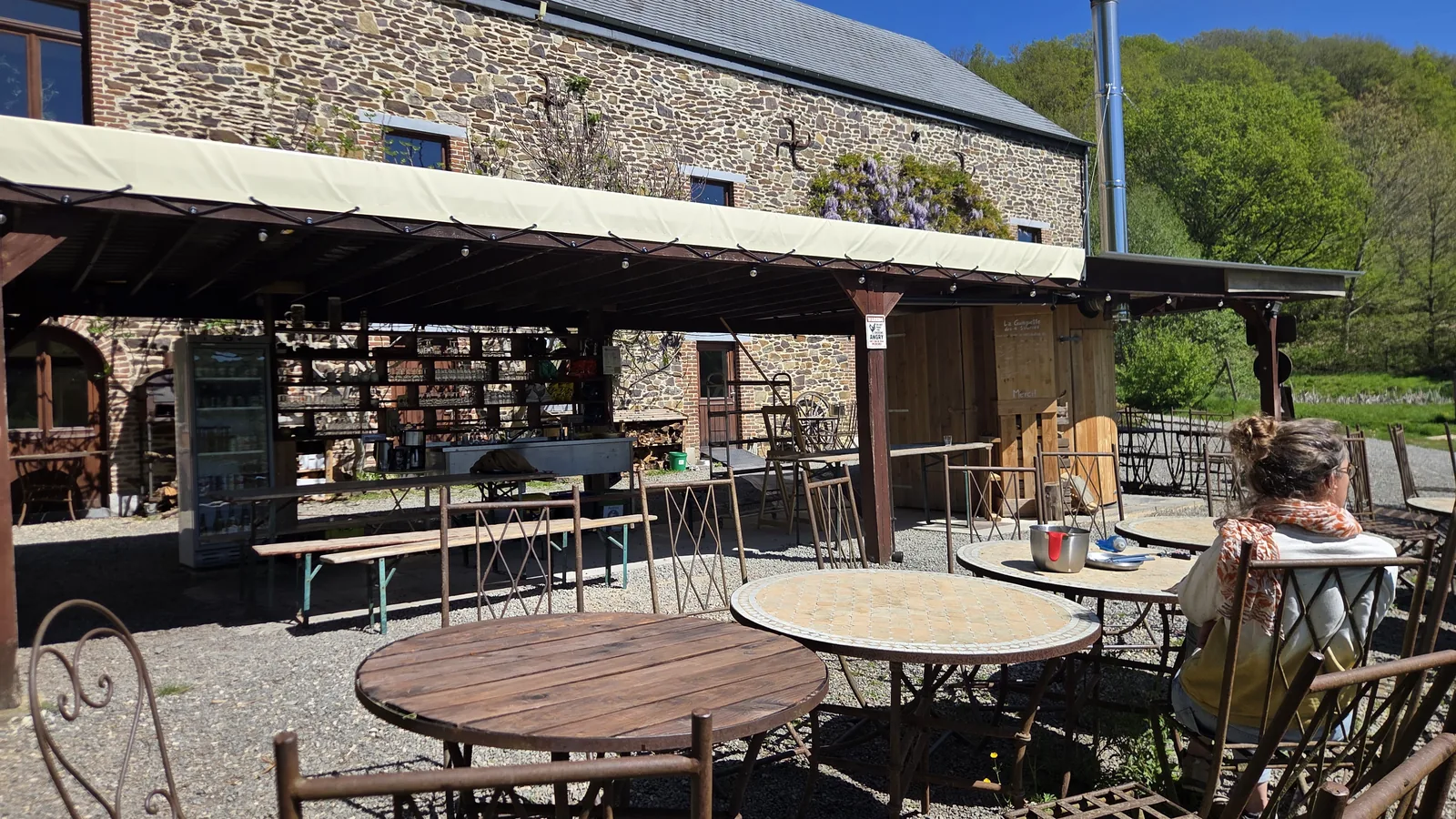 Terrasse extérieure des 4 Sources sous un grand auvent en toile, plusieurs tables rondes et chaises en fer forgé sur le gravier, mur en pierre du bâtiment principal en arrière-plan