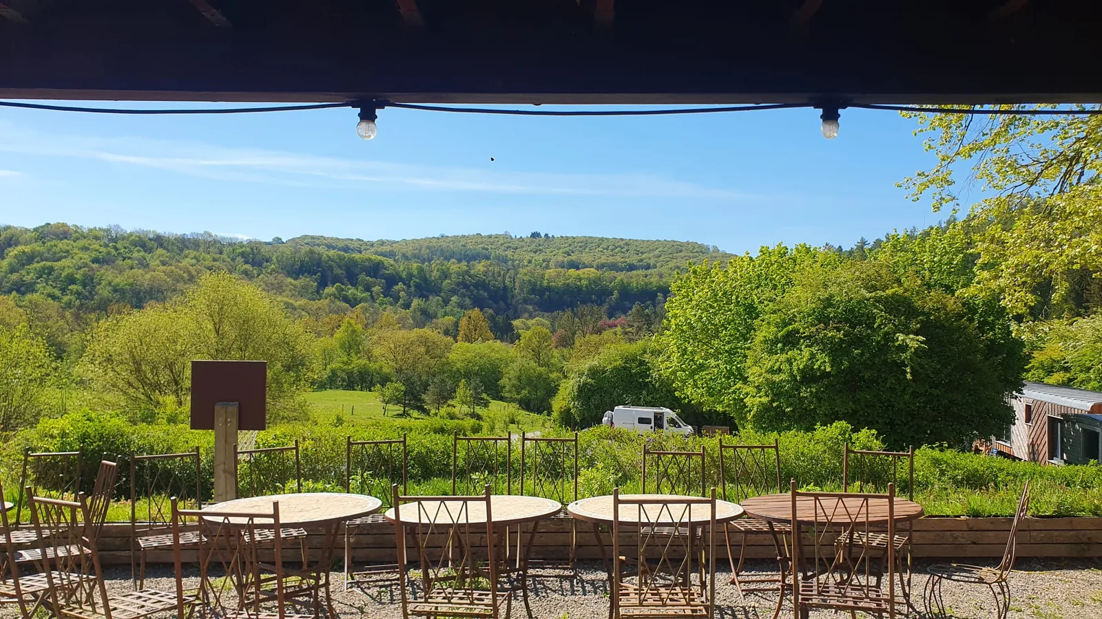 Vue panoramique sur la vallée d'Yvoir prise depuis la terrasse couverte du bâtiment principal, tables rondes et chaises en fer forgé au premier plan, sentier qui descend vers la prairie, collines boisées au fond