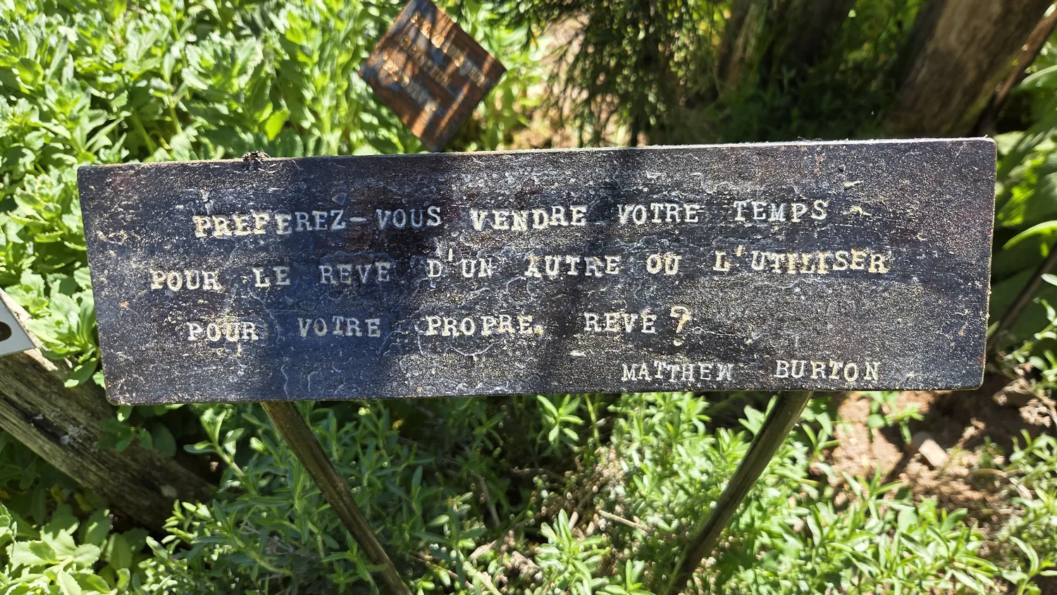 Petite plaque gravée posée dans la végétation du jardin : « Préférez-vous vendre votre temps pour le rêve d'un autre ou l'utiliser pour votre propre rêve ? Matthew Burton »