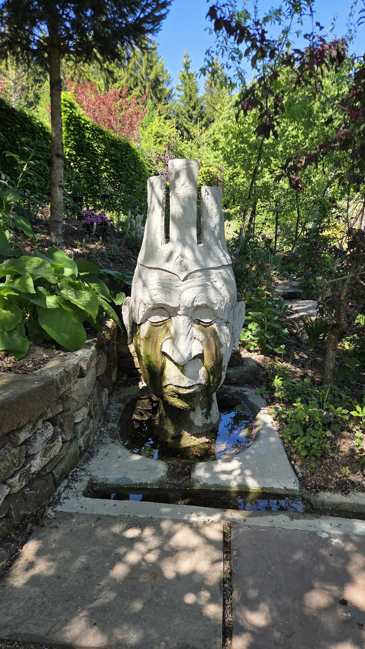 Sculpture en pierre d'un visage féminin serein, posée sur un socle au milieu de la végétation du jardin