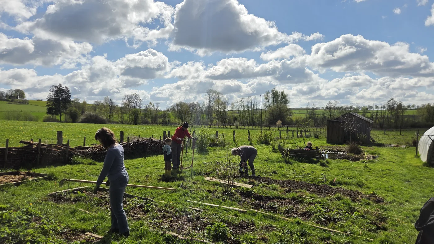 Plusieurs adultes et enfants au travail dans le potager : préparation de planches, plantation, palissages tendus, ciel bleu et nuages