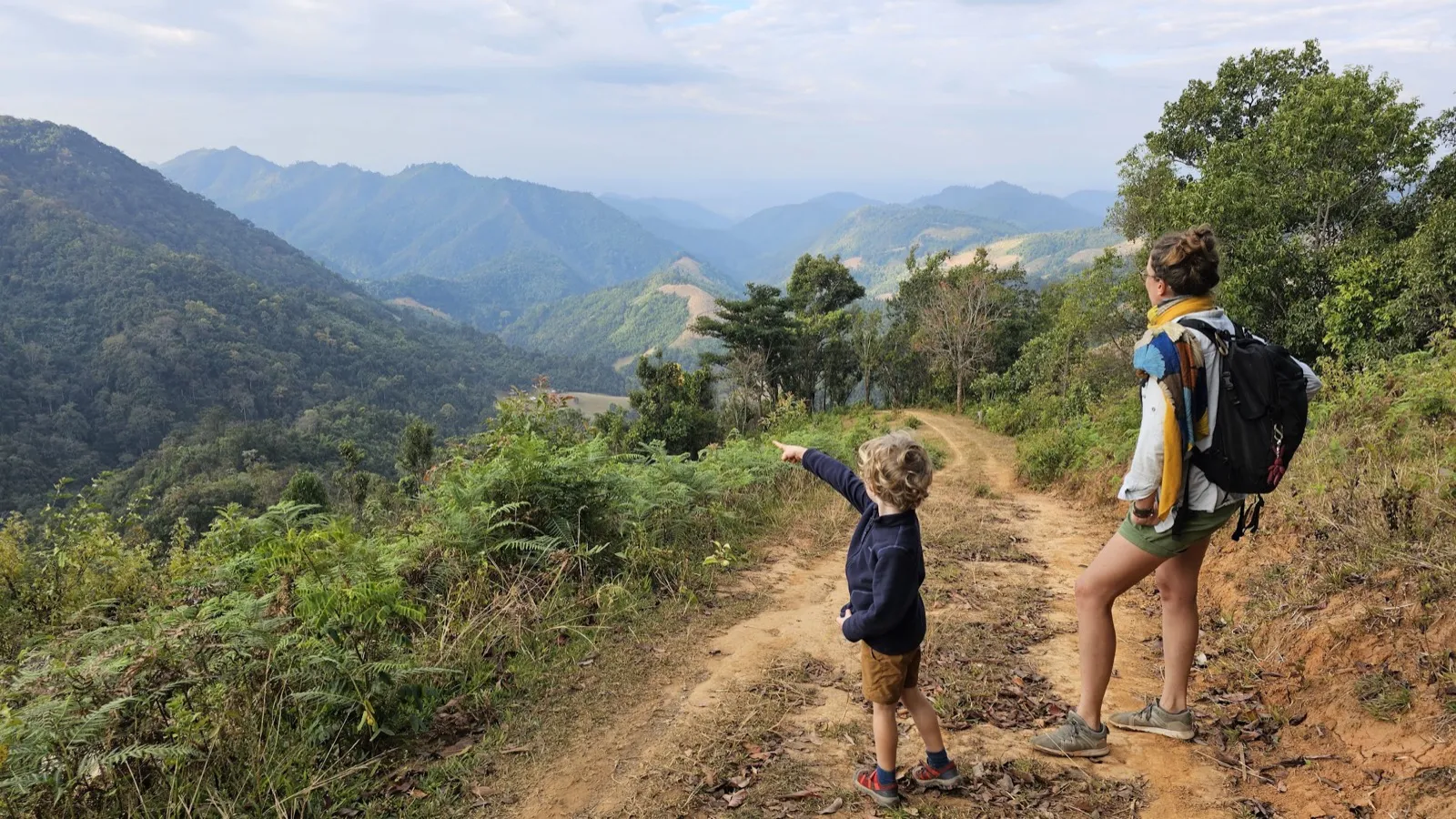Un enfant aux cheveux bouclés pointe du doigt un panorama de montagnes verdoyantes sur un sentier de terre rouge, accompagné d'une femme avec un sac à dos, en Asie du Sud-Est