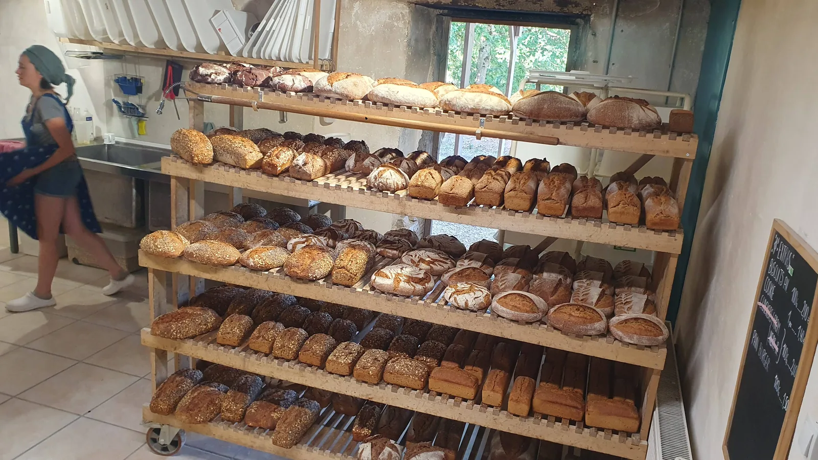 L'échelle à pain sur roulettes, ses cinq niveaux chargés de miches et de pains aux graines, Tania au fond du fournil